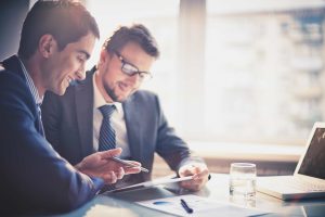 Two men in business suits collaborating on a tablet by an office window.
