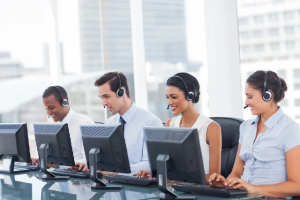 A smiling row of young men and women wearing headsets and working at computers.