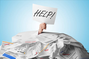 A hand sticking out of a pile of papers and holding up a paper with 'HELP!' scribbled in marker.